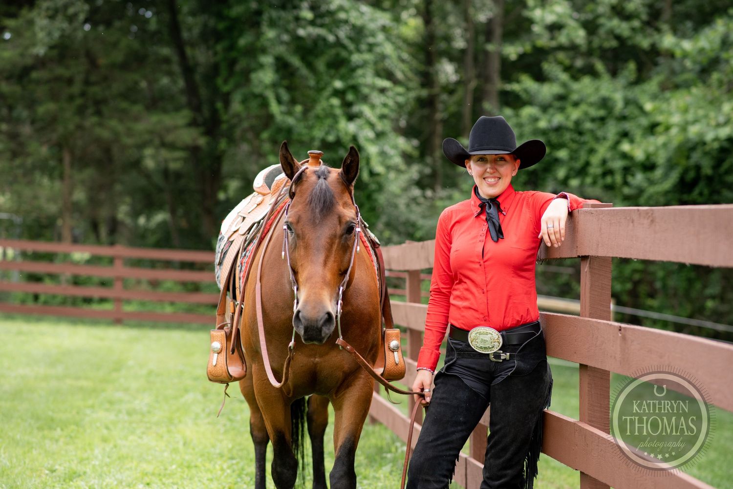 Equestrian in red shirt and black cowboy hat stands with horse by wooden fence.