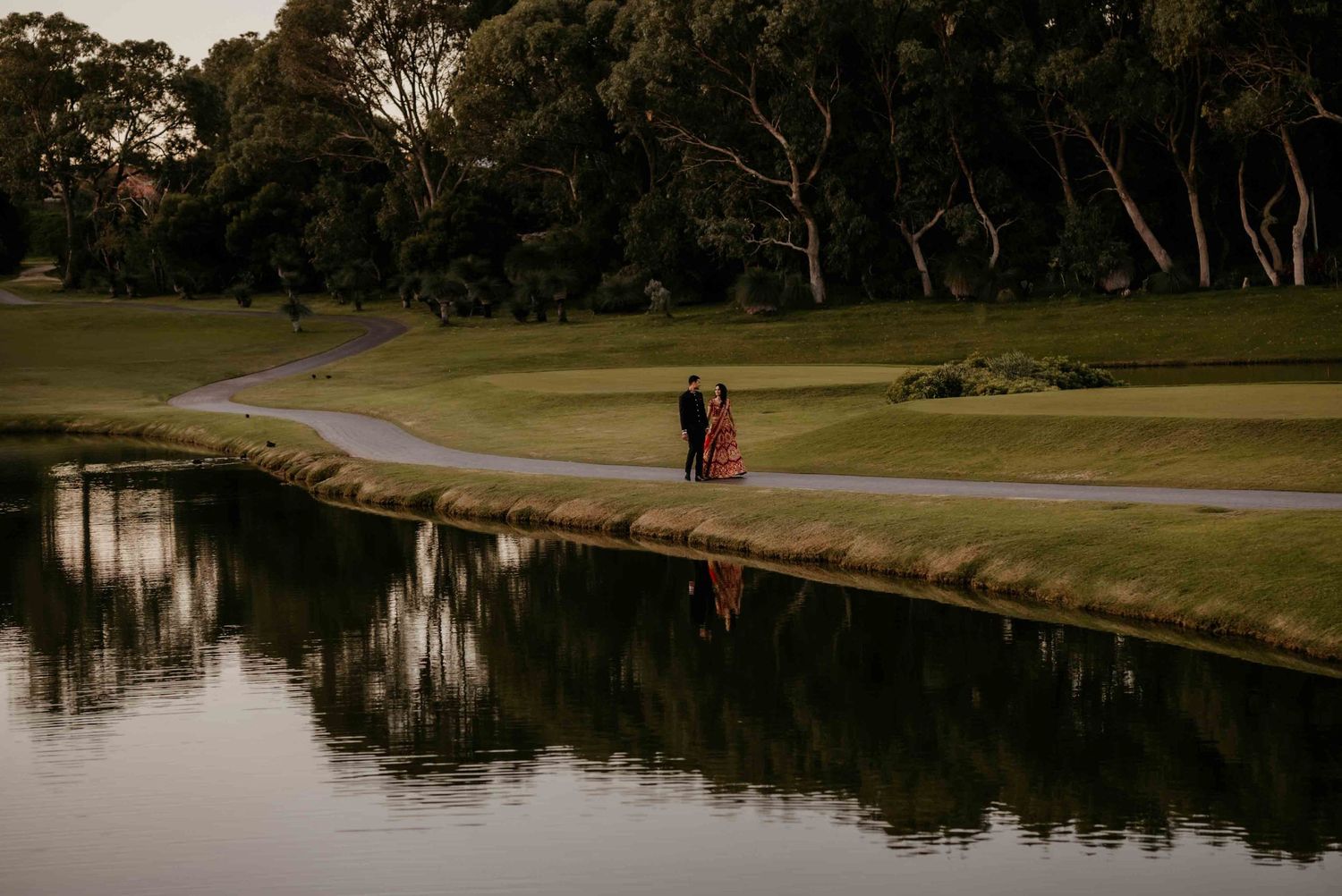Two silhouettes walk along a winding path beside a reflective pond at dusk with trees in the background.