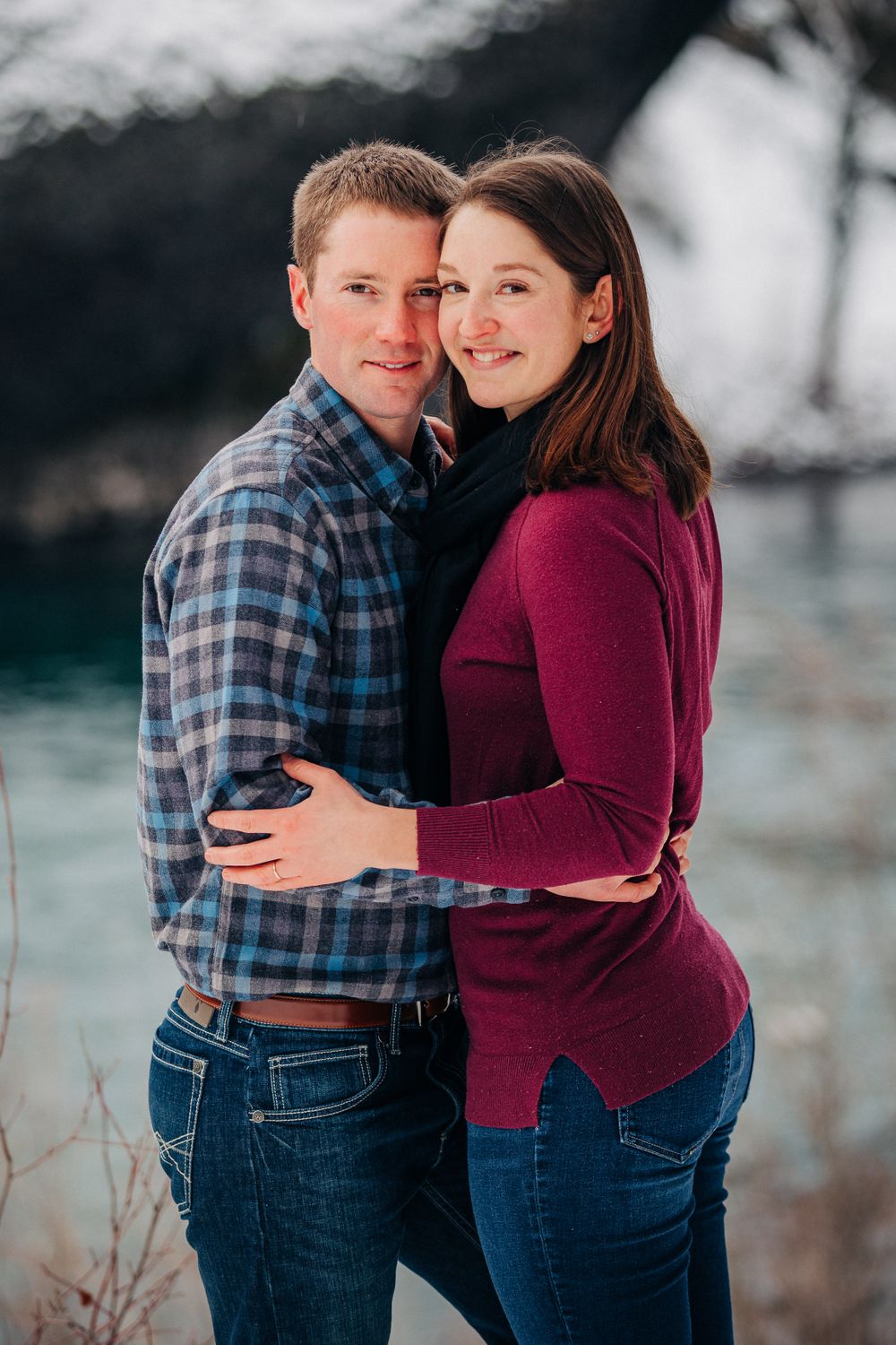A couple in casual winter attire embrace against a snowy mountain backdrop.
