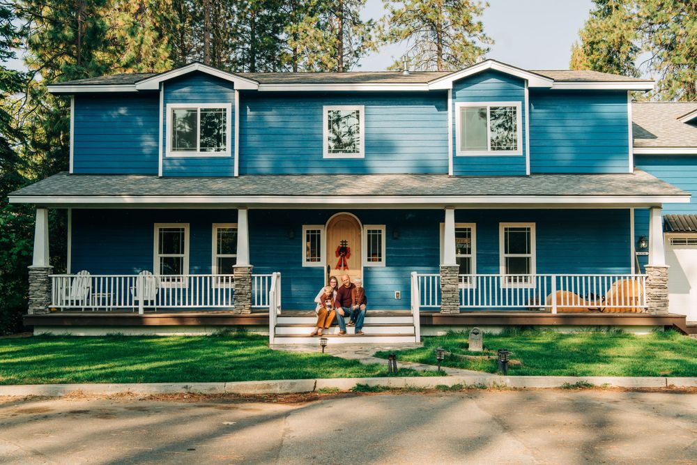 A blue two-story country house with white trim and wraparound porch nestled among tall pine trees.