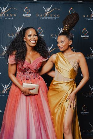 Two elegantly dressed attendees in coral and gold evening gowns pose at formal event.
