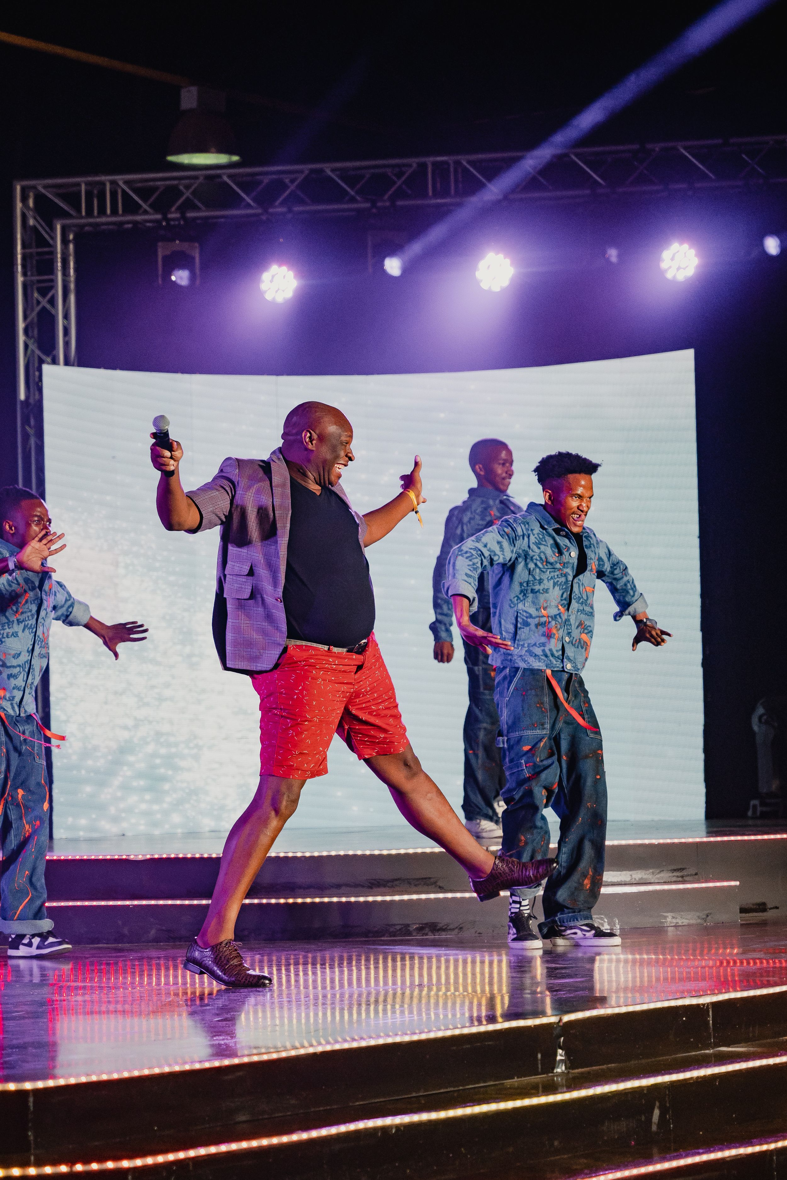 Performers dancing energetically on stage under purple spotlights with a white backdrop.