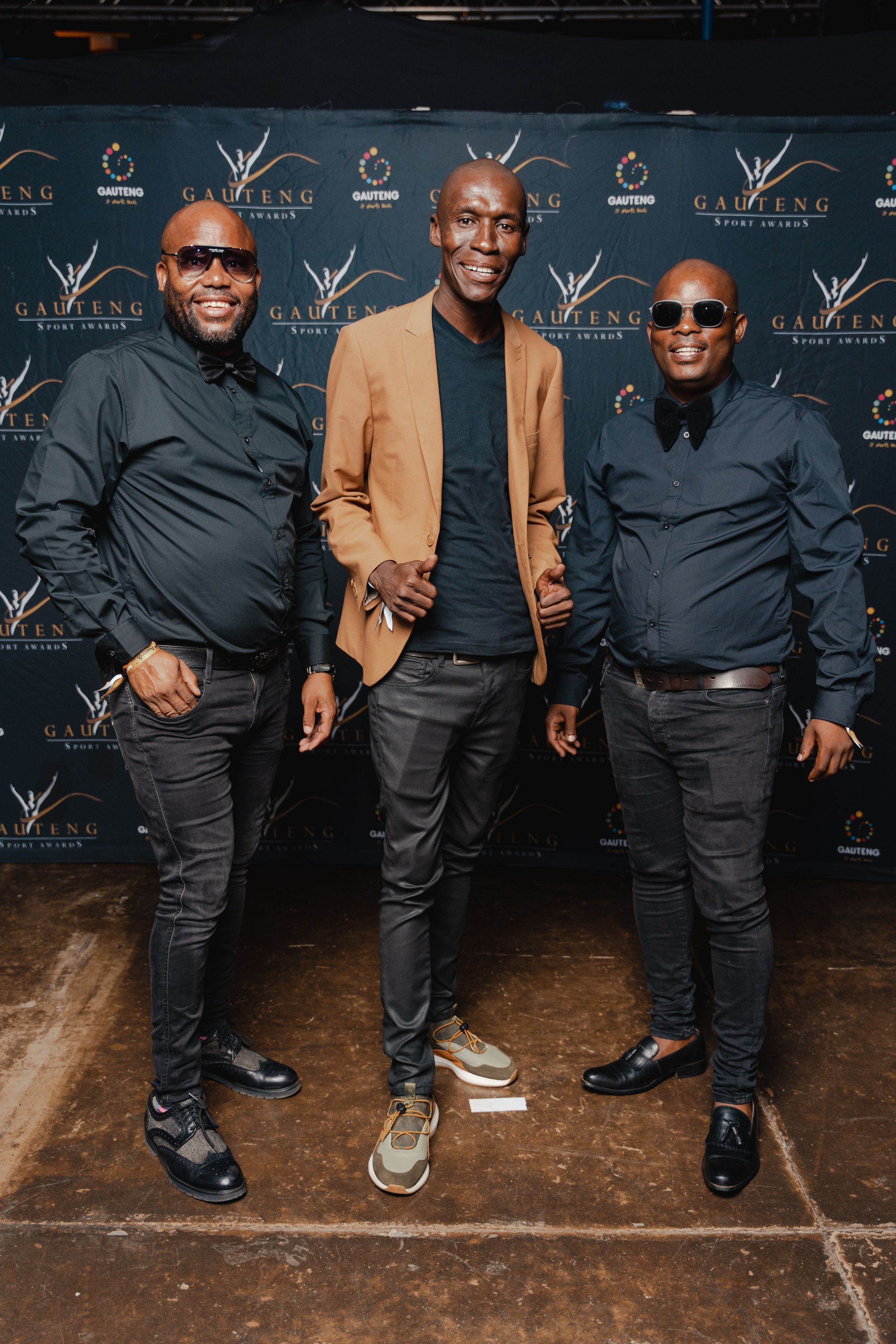 Three men in stylish attire pose against a branded step-and-repeat wall at an event.