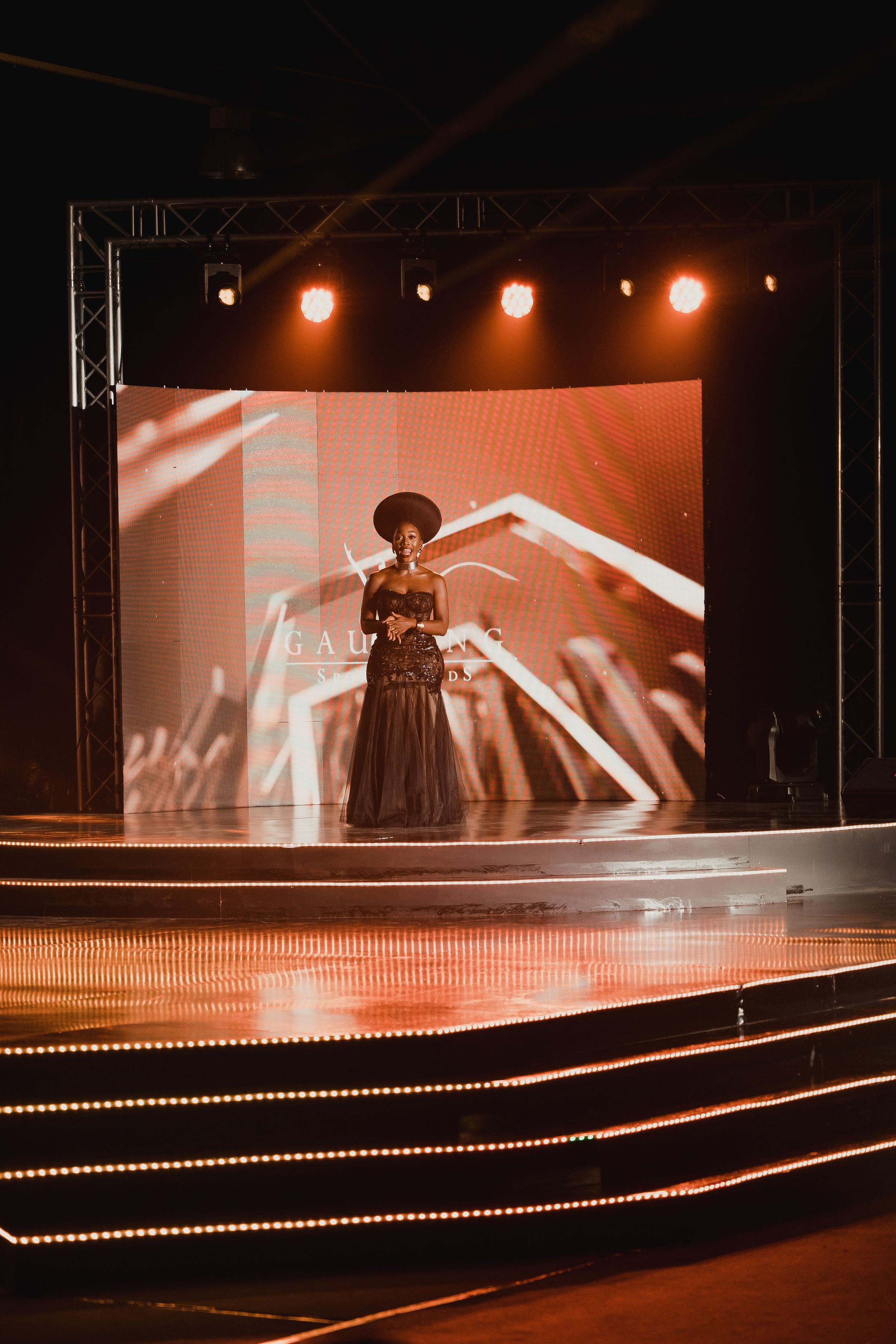 Dramatic stage performance with a performer in a black dress and hat silhouetted against orange lighting and geometric backdrop.