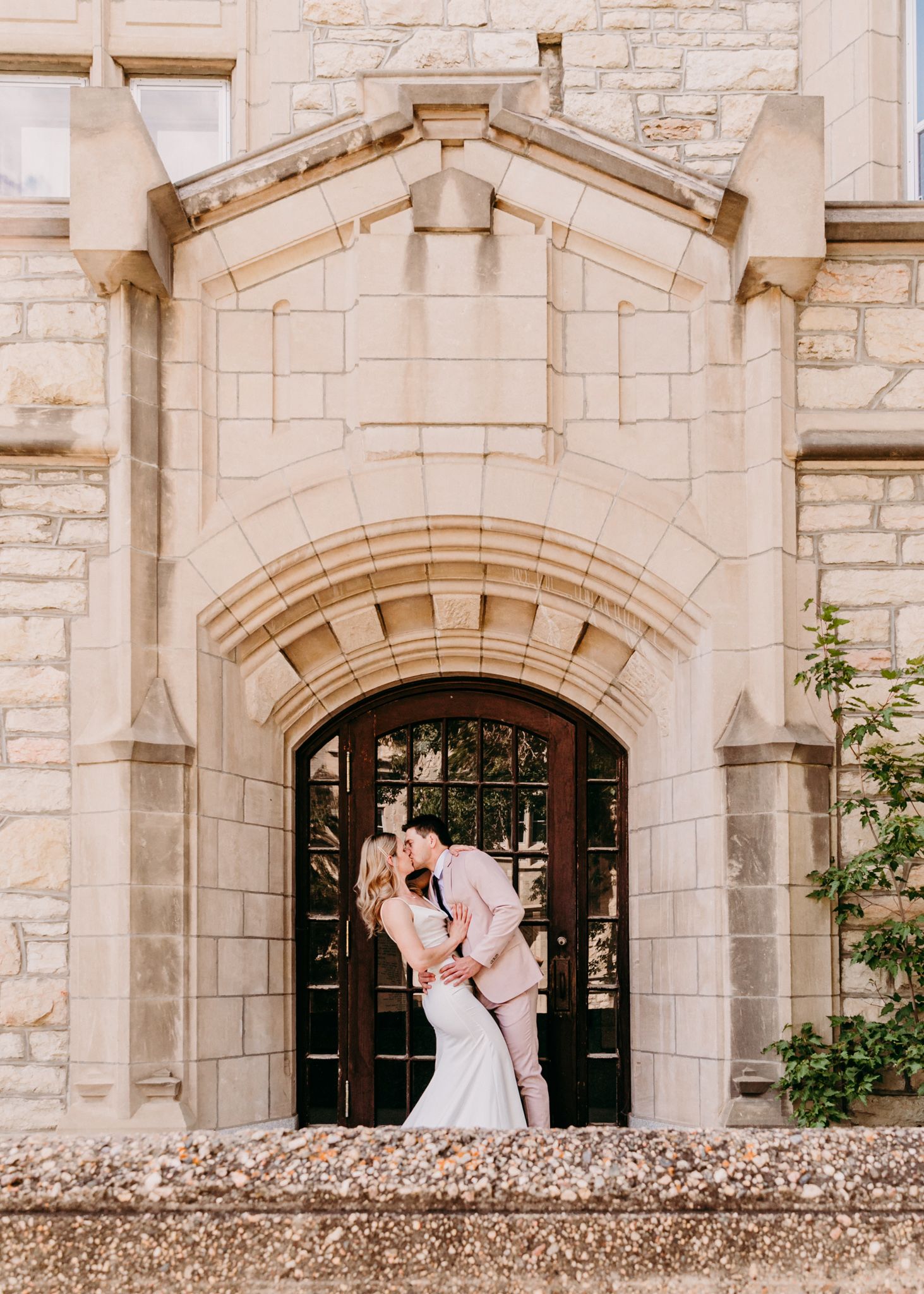 Romantic wedding couple sharing a kiss under a grand stone archway of a historic building.