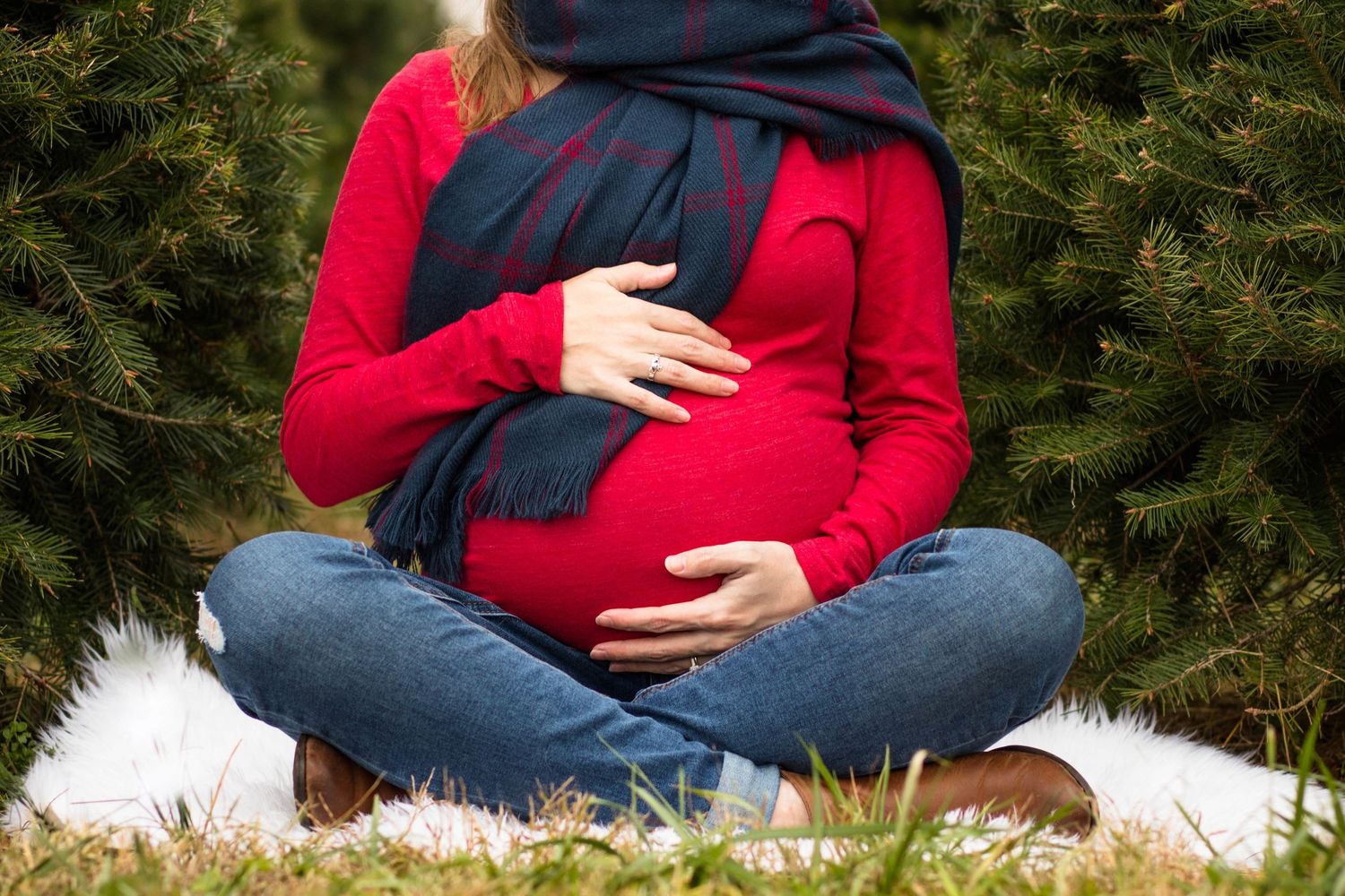 Person in red sweater and jeans showing pregnant belly against evergreen bushes.