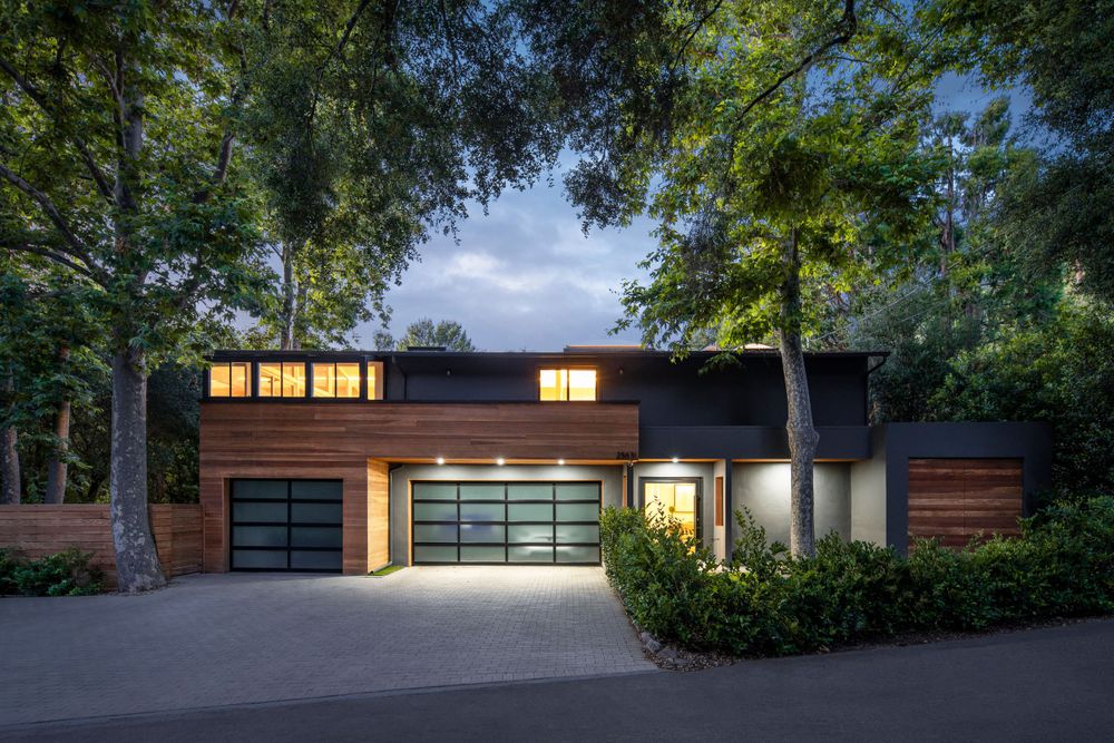 Modern wood and black home with illuminated garage doors at dusk.