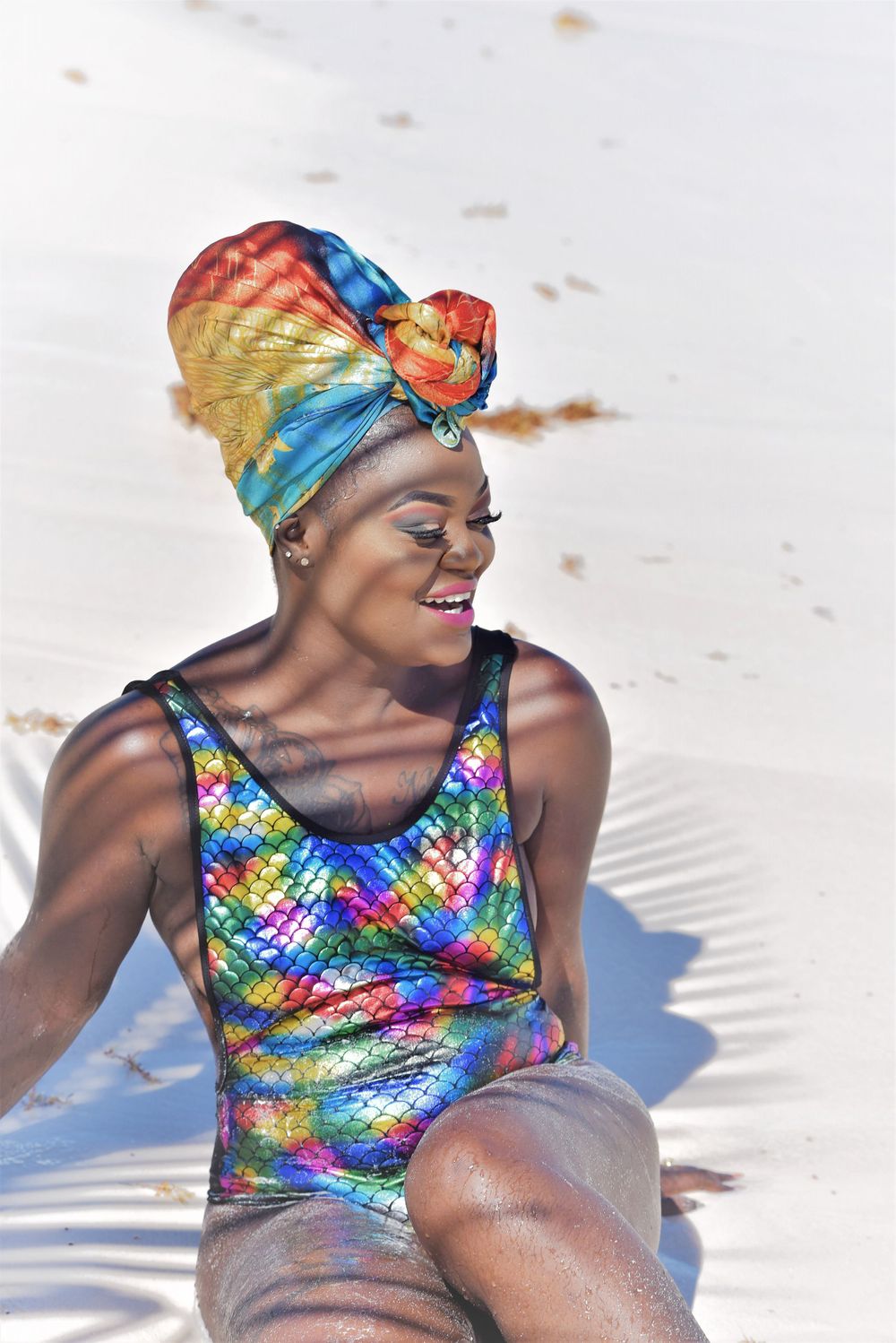 Woman in colorful turban and sequined swimsuit sitting on white sand beach.