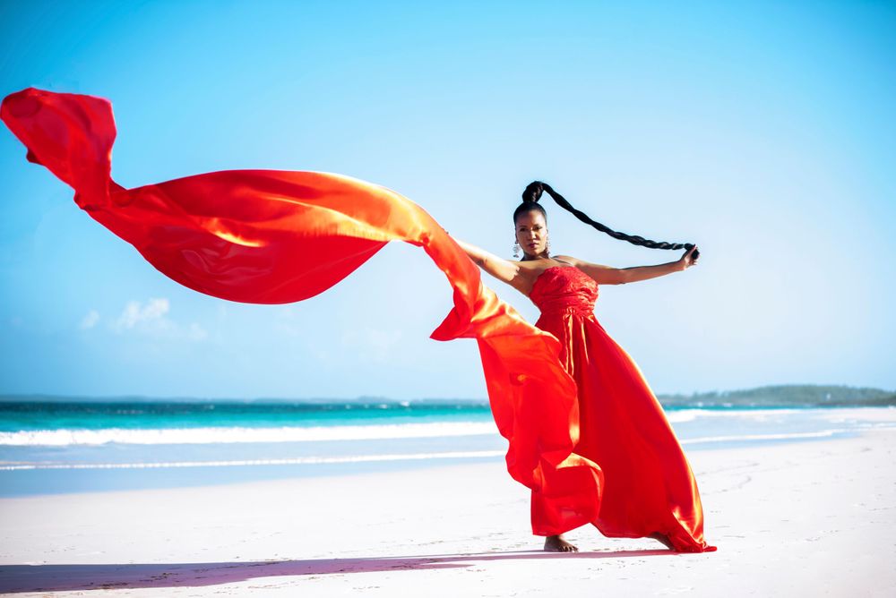 Dramatic fashion shot of dancer in flowing red dress on beach with billowing fabric against blue sky.