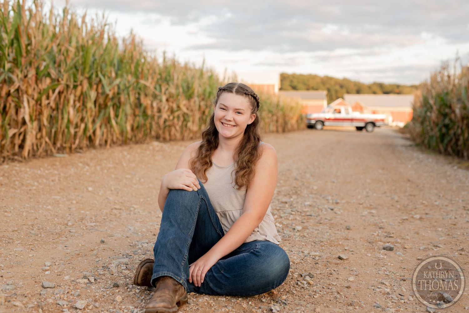 Sitting cross-legged on a rural path with cornfields and barns in the background.