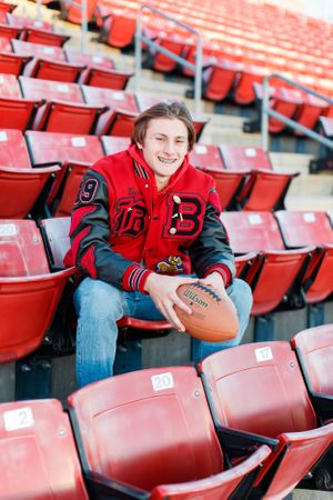 Senior boy wearing red varsity jacket sits with football in empty red stadium seats for senior session in Springfield, MO.