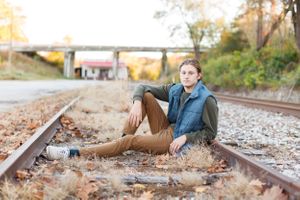 Senior boy in casual attire sitting on railroad tracks surrounded by fallen autumn leaves at golden hour for a senior session in Springfield