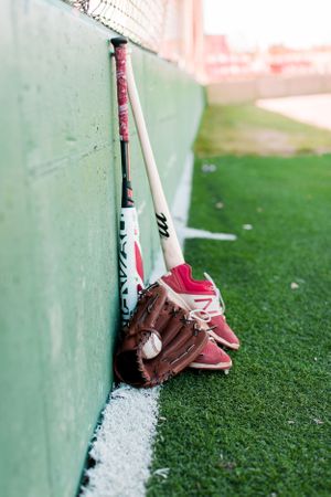 Baseball equipment including bats, glove, and cleats leaning against green wall on baseball field.