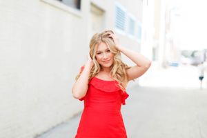 Senior girl in a vibrant red dress poses confidently against a light colored urban wall for a senior session in Springfield, MO..