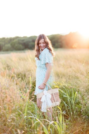 Senior girl wearing light blue dress carrying wicker basket in tall grass field during golden hour for a senior session in Springfield, MO.