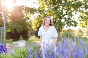 Senior girl in white dress standing among blooming purple delphiniums in a garden at sunset for a senior session in Springfield, MO.