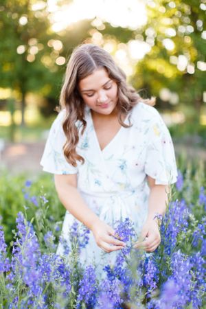 Senior girl in white floral dress gently touching purple delphinium flowers in sunlit garden for a senior session in Springfield, MO.
