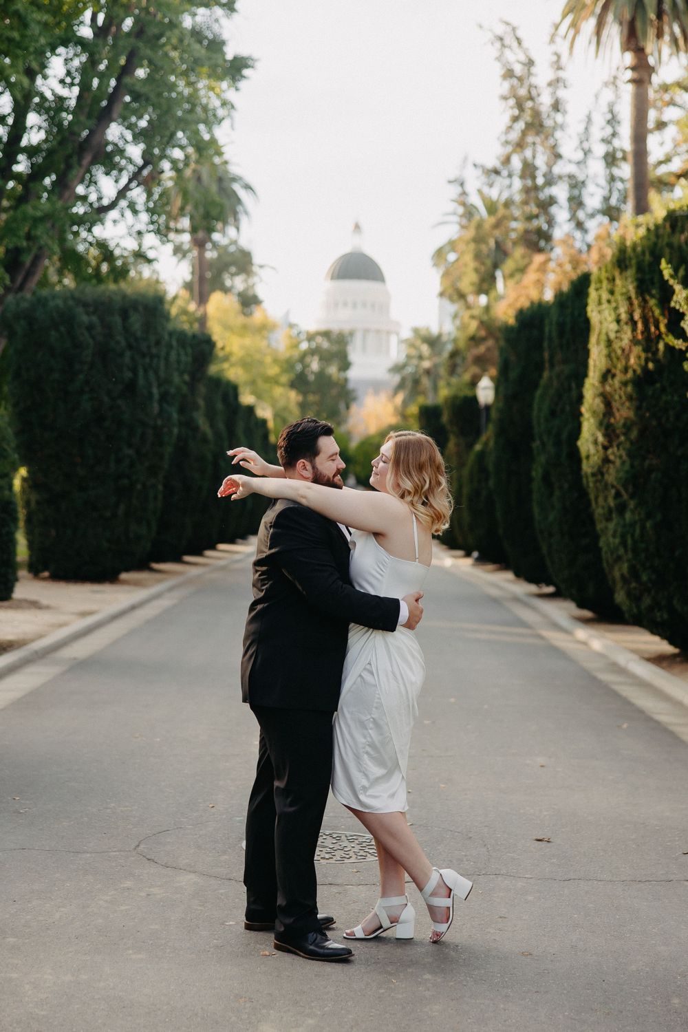 A couple in formal attire dancing together on a tree-lined path with a domed building in the background.