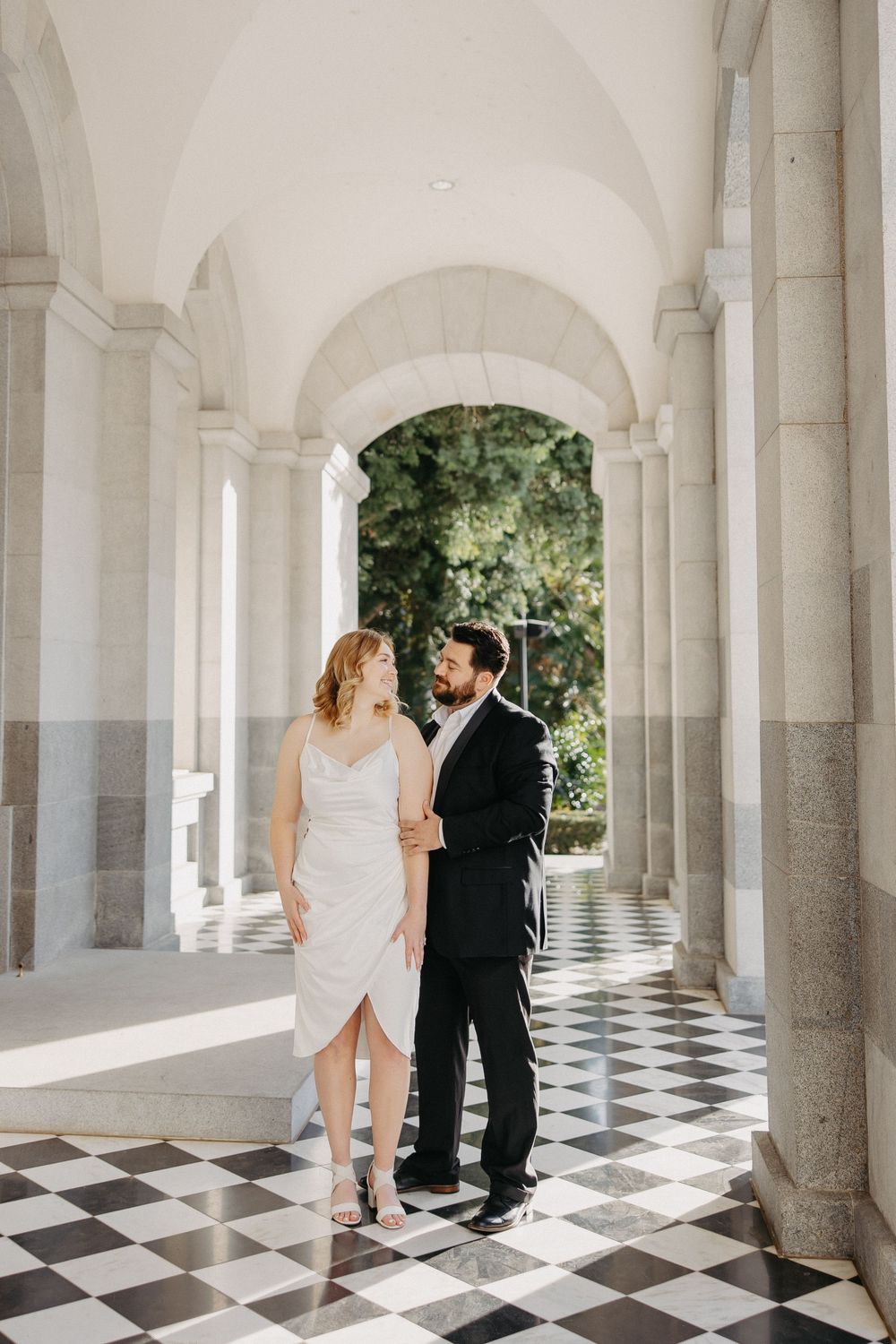 Romantic couple poses beneath ornate white columns on a black and white checkered floor.