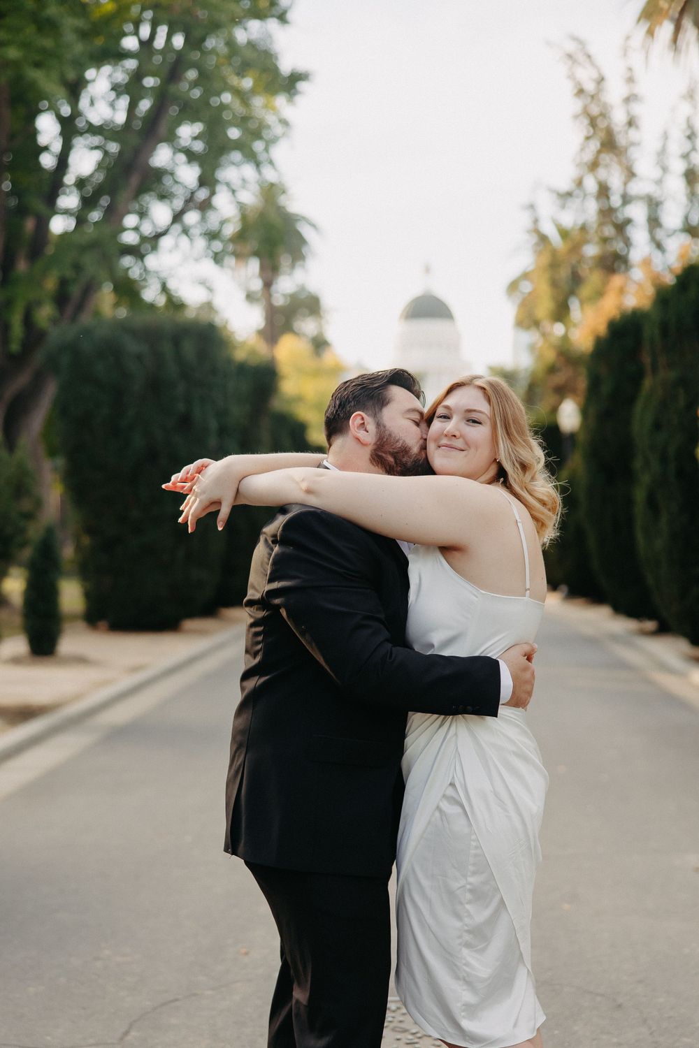 A romantic embrace on a palm-lined walkway with a domed structure visible in the distance.