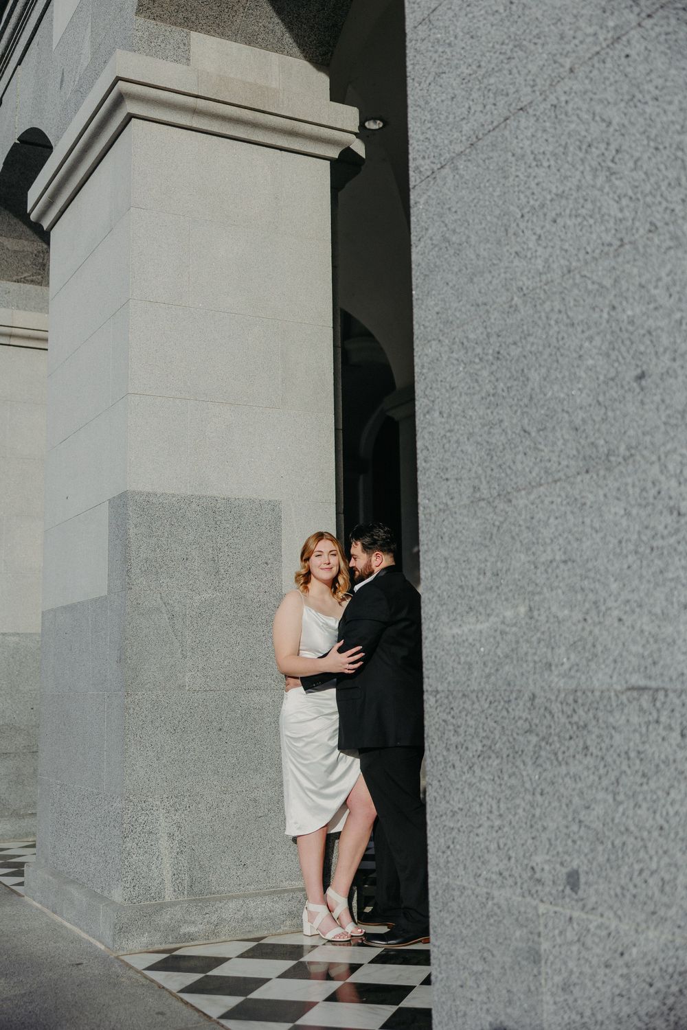 Couple shares tender moment against stone pillar with checkerboard floor beneath.