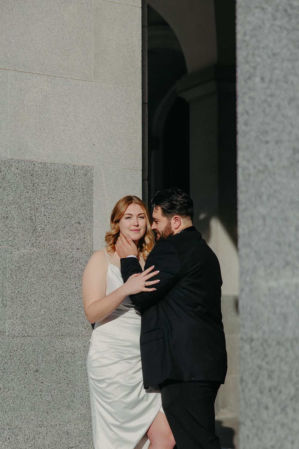 Intimate embrace between couple against stone column in formal wedding attire.