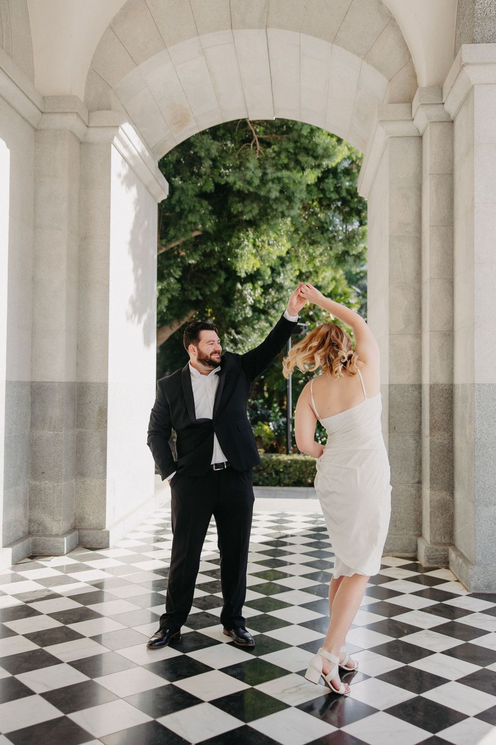 A couple dances elegantly under a classical archway with checkered floor tiles.