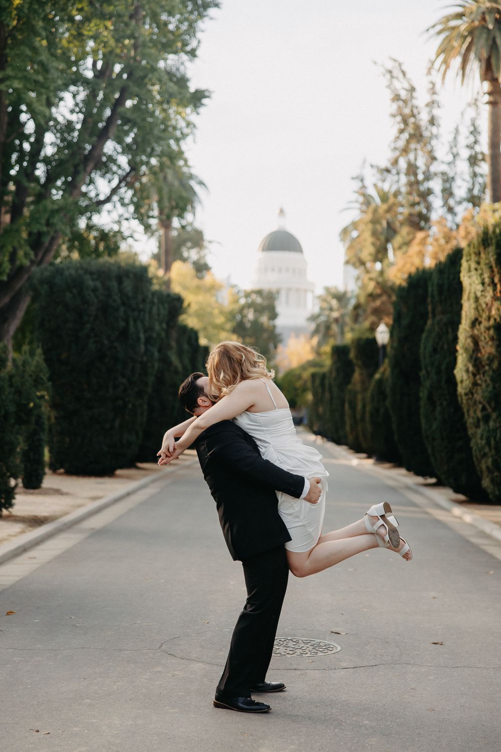 Joyful lift pose between couple on tree-lined pathway with domed building in background.