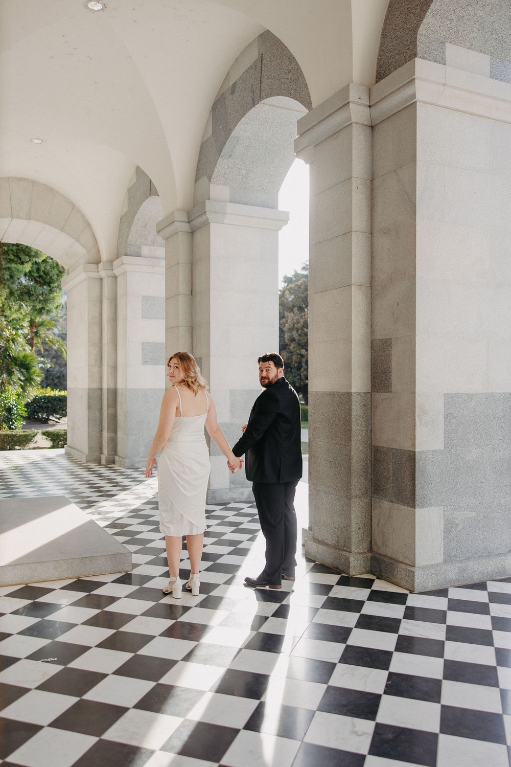 Couple walks through sunlit classical colonnade with black and white checkerboard flooring.
