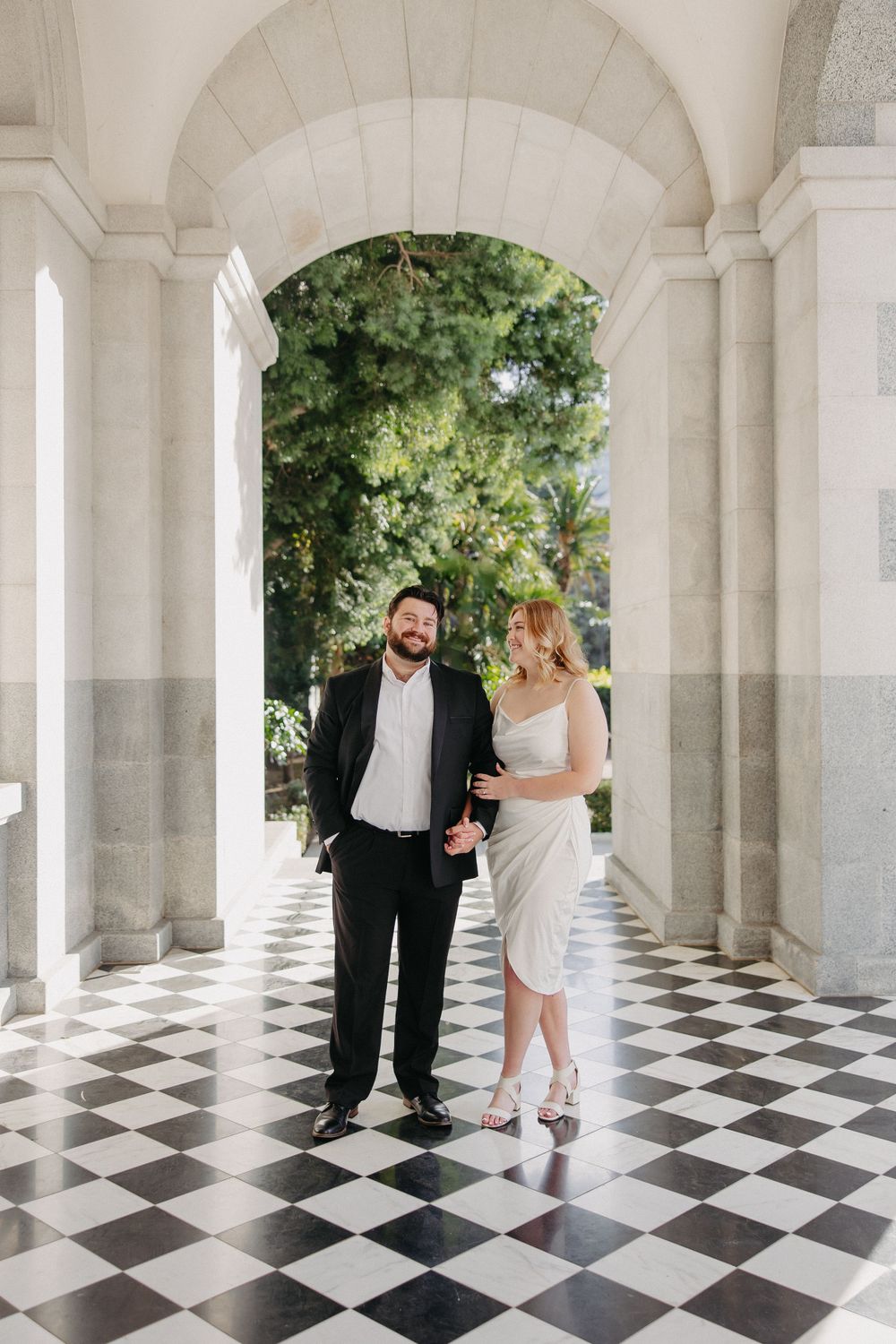 A couple stands in a grand colonnade with checkered floors and arched architecture.