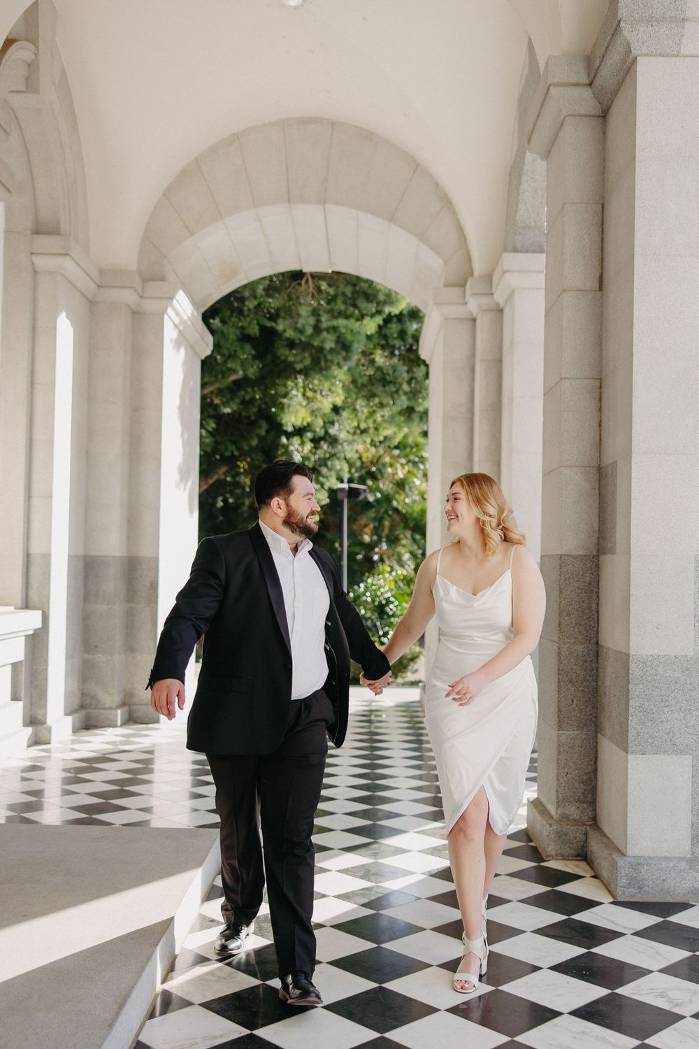 A couple walks together on a checkered marble floor beneath elegant white archways with greenery visible outside.