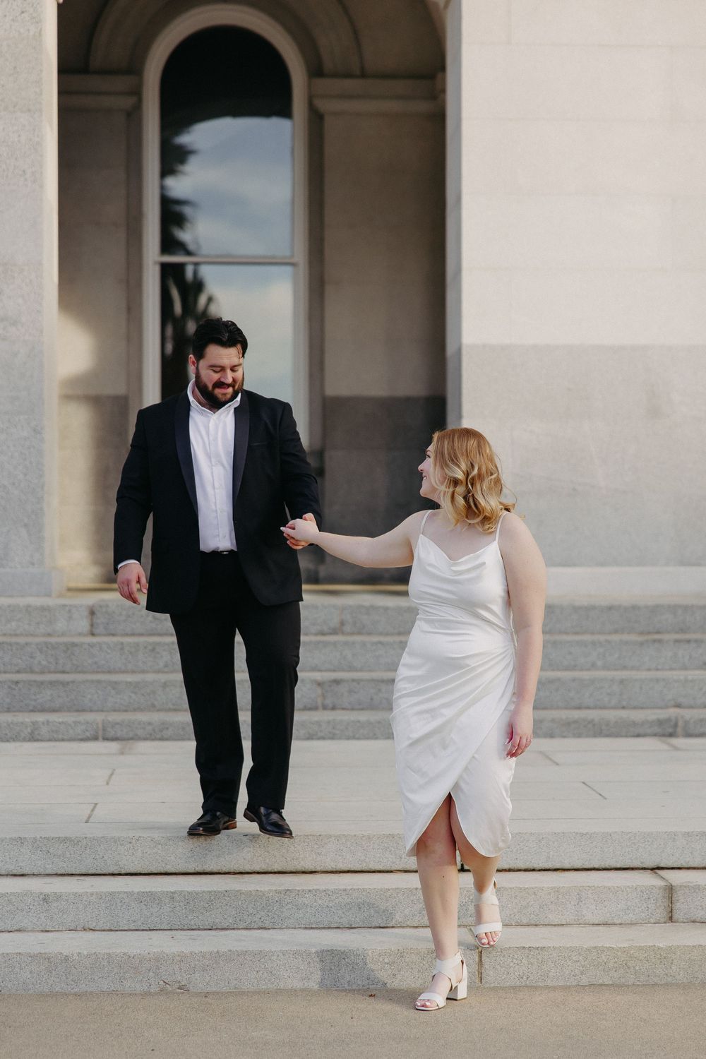 Couple in formal attire holding hands on stone steps of classical building.