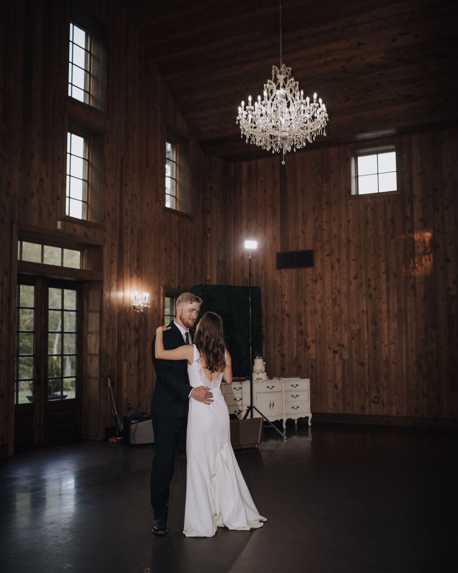 Couple shares intimate dance in rustic wooden venue under crystal chandelier.