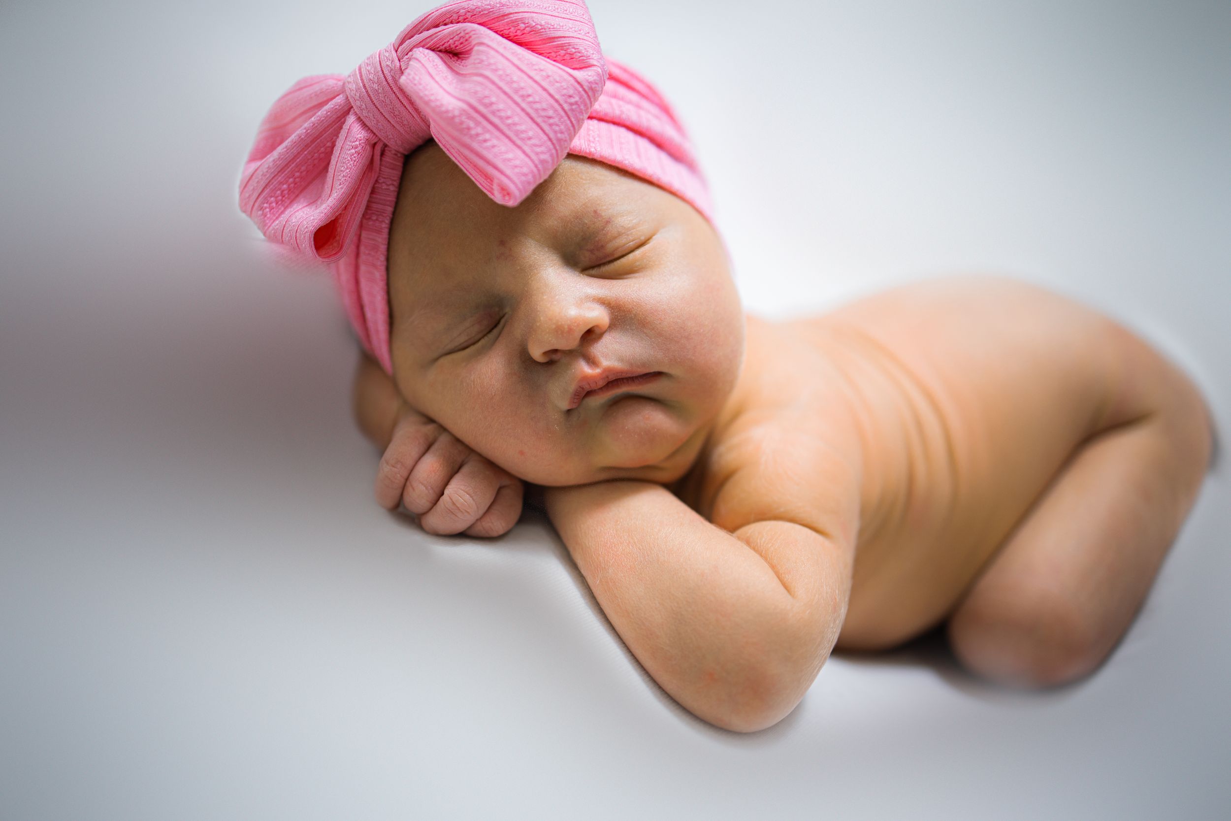 Sleeping newborn wearing pink bow headband on white background.