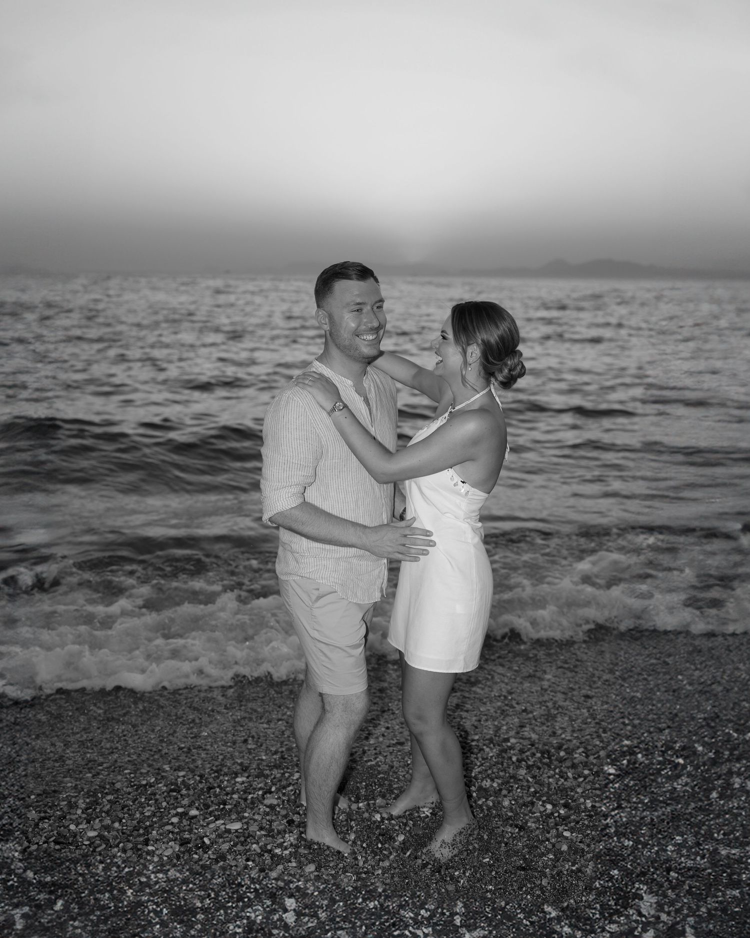 A couple embraces on a beach at sunset in black and white.