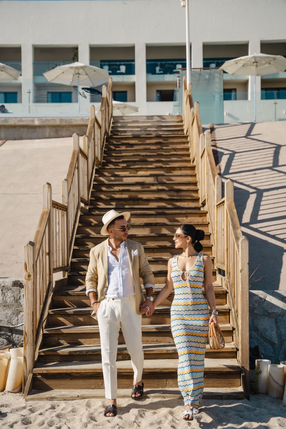 Stylish couple descending wooden beach stairs in summer resort wear.
