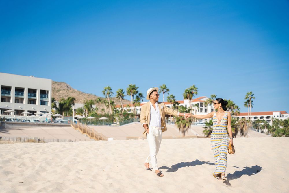 A couple walking barefoot on a sunny beach with palm trees and buildings in the background.