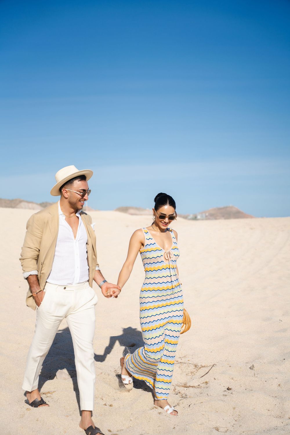 A stylish couple in summer attire holding hands while walking through desert sand dunes.