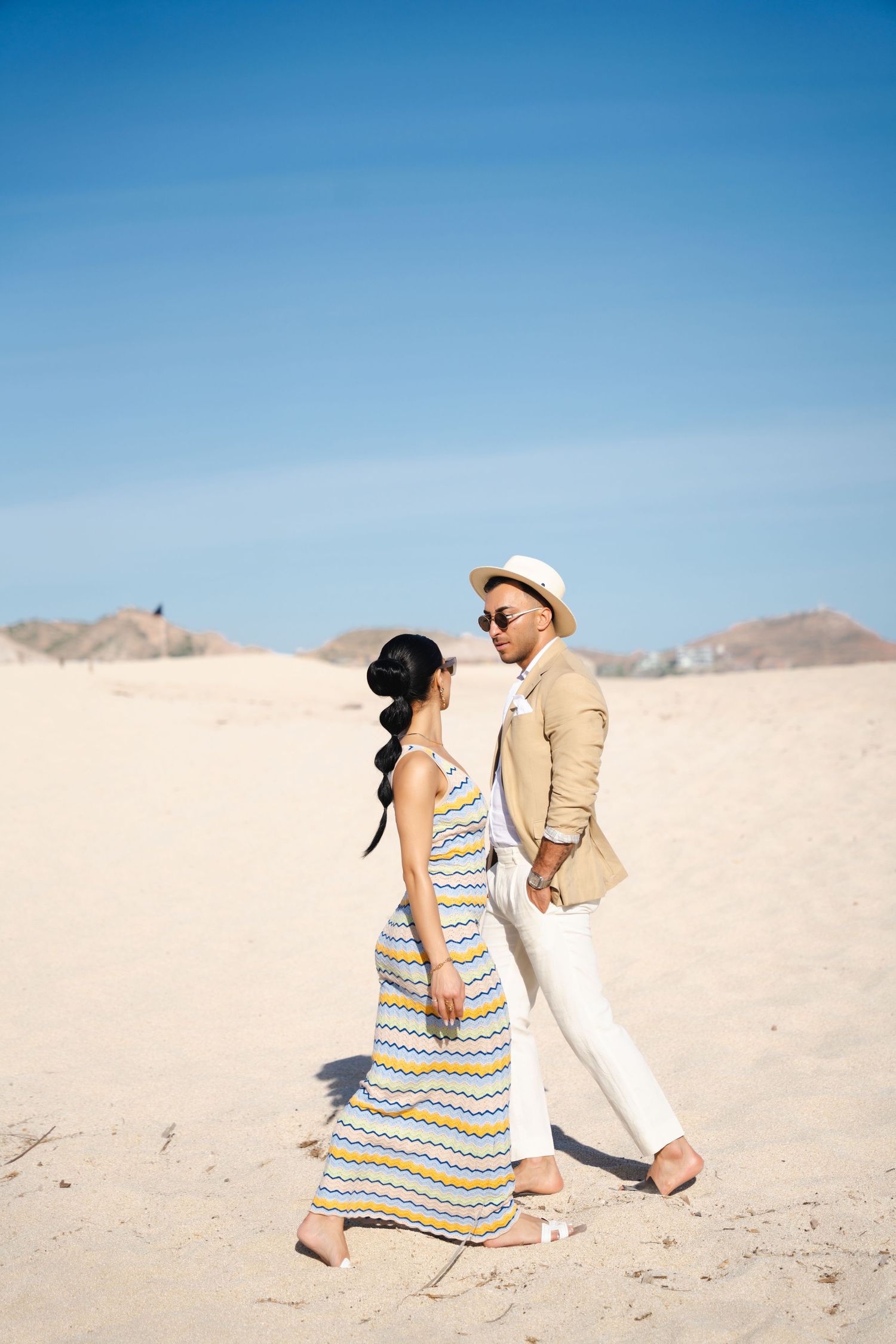 Couple in coordinated beachwear walking together on sandy dunes under bright blue sky.