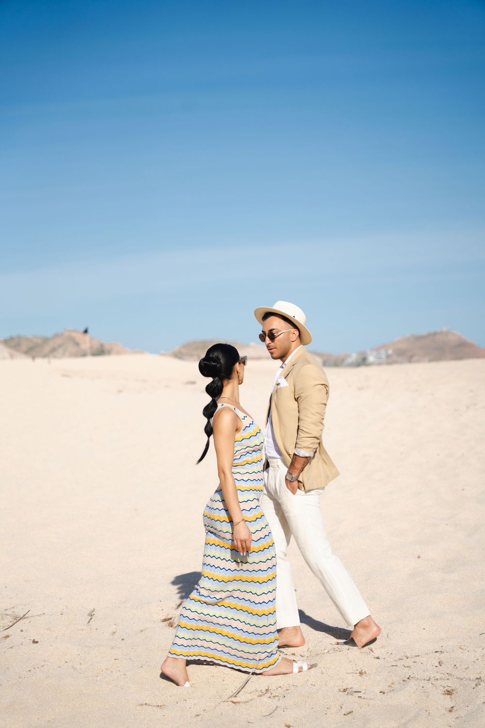 Couple in coordinated beachwear walking together on sandy dunes under bright blue sky.