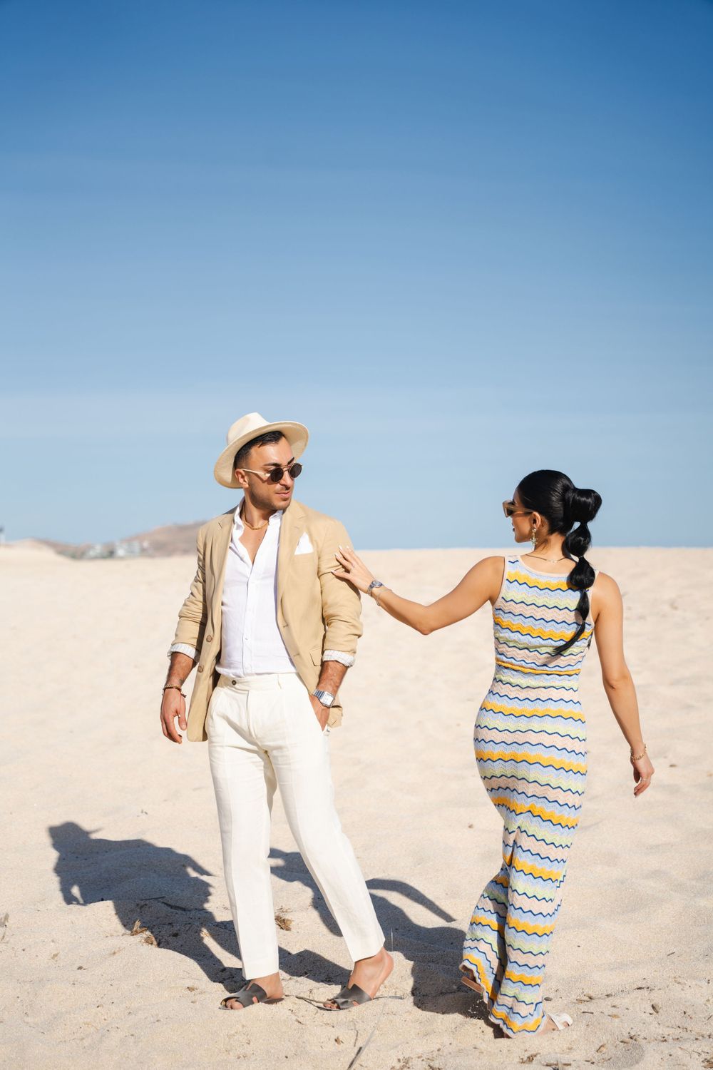 Couple in casual beach attire walking hand in hand across desert sand dunes.