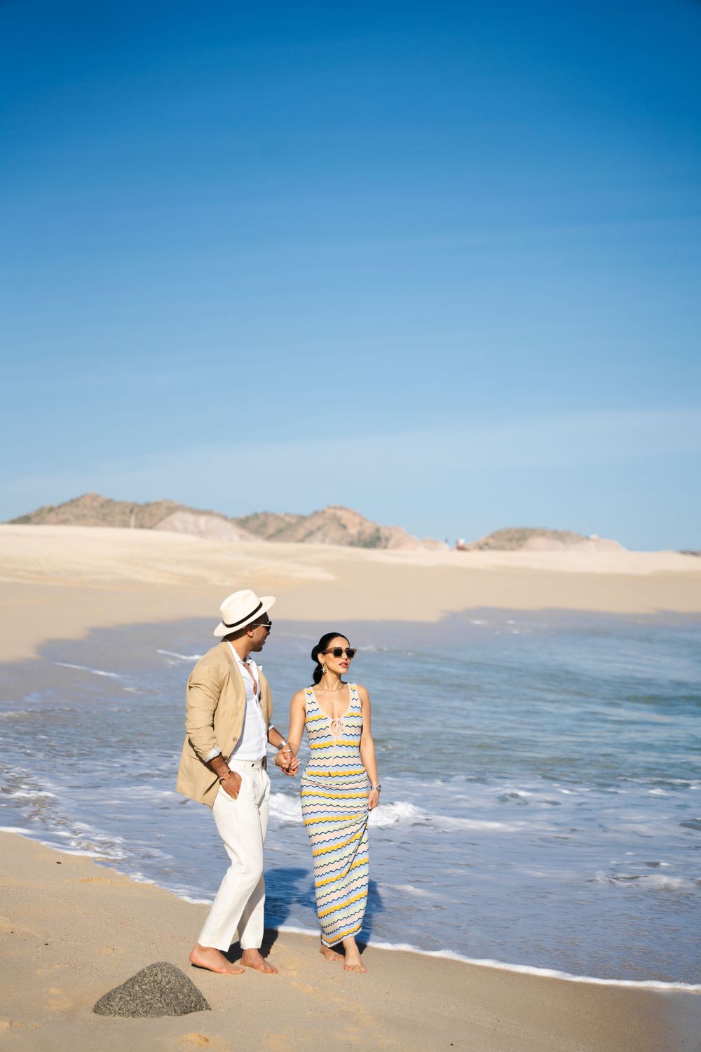 Couple in beachwear walking along ocean shoreline with mountains in background.