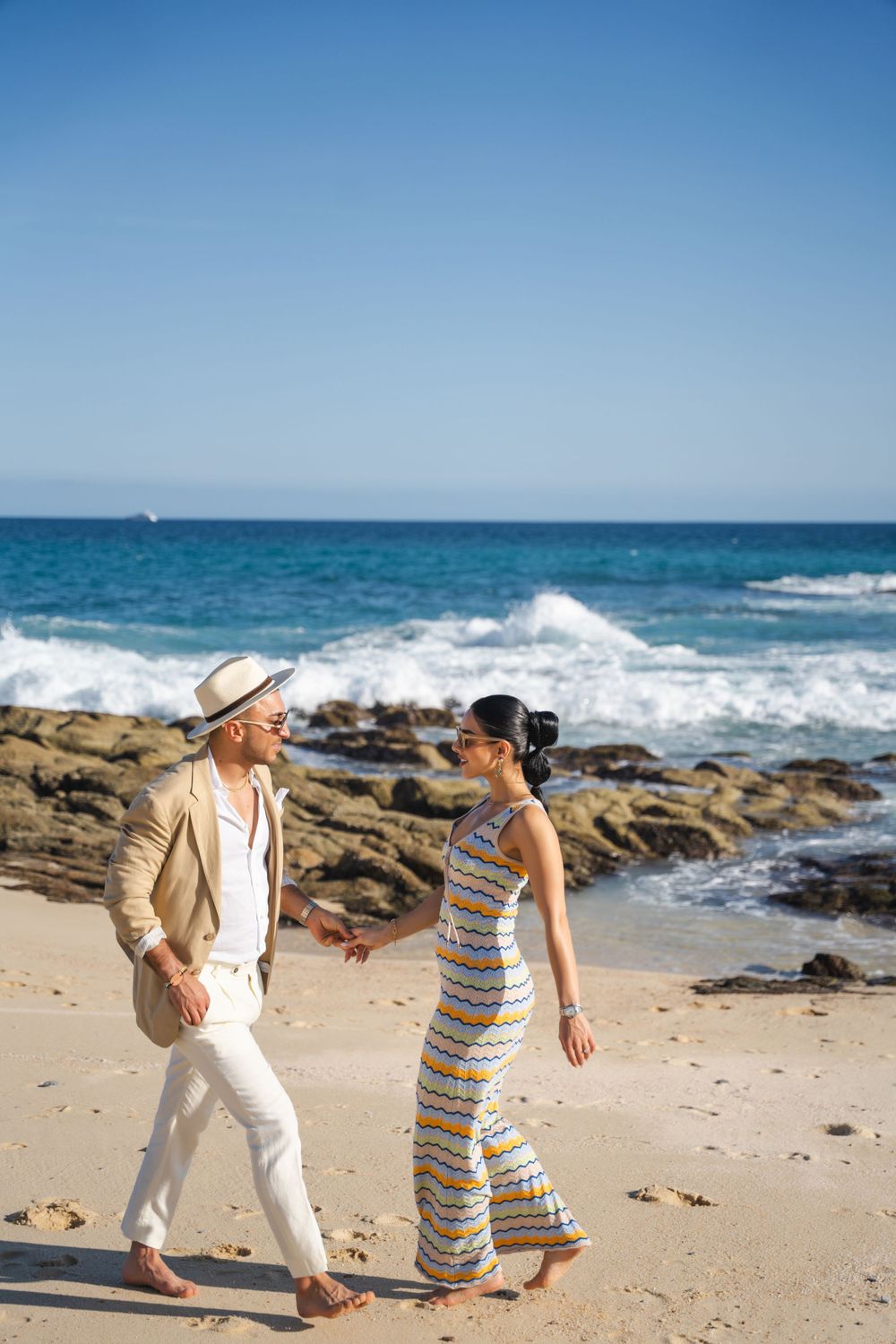Couple in coordinated outfits walking on rocky beach with crashing waves.