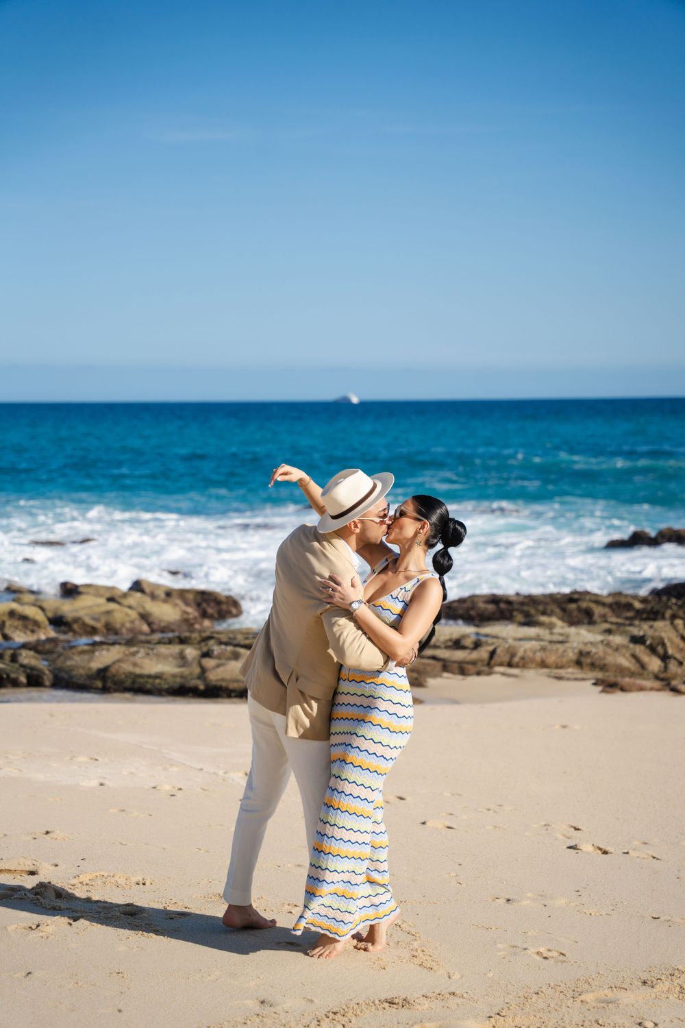Romantic couple embracing on beach with ocean waves and rocky coastline in background.