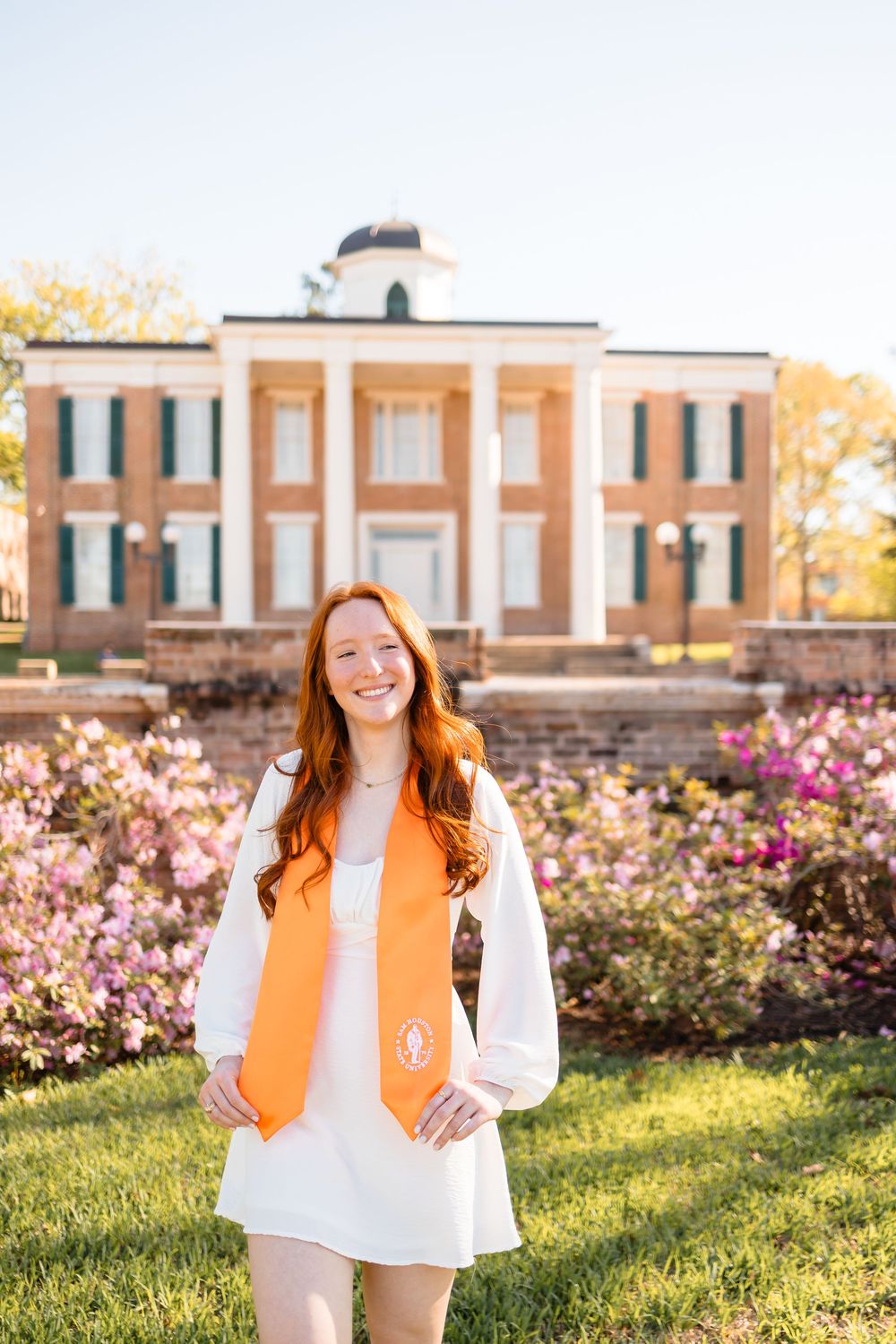 Smiling redhead graduate in white dress with orange stole poses before blooming flowers and brick building.