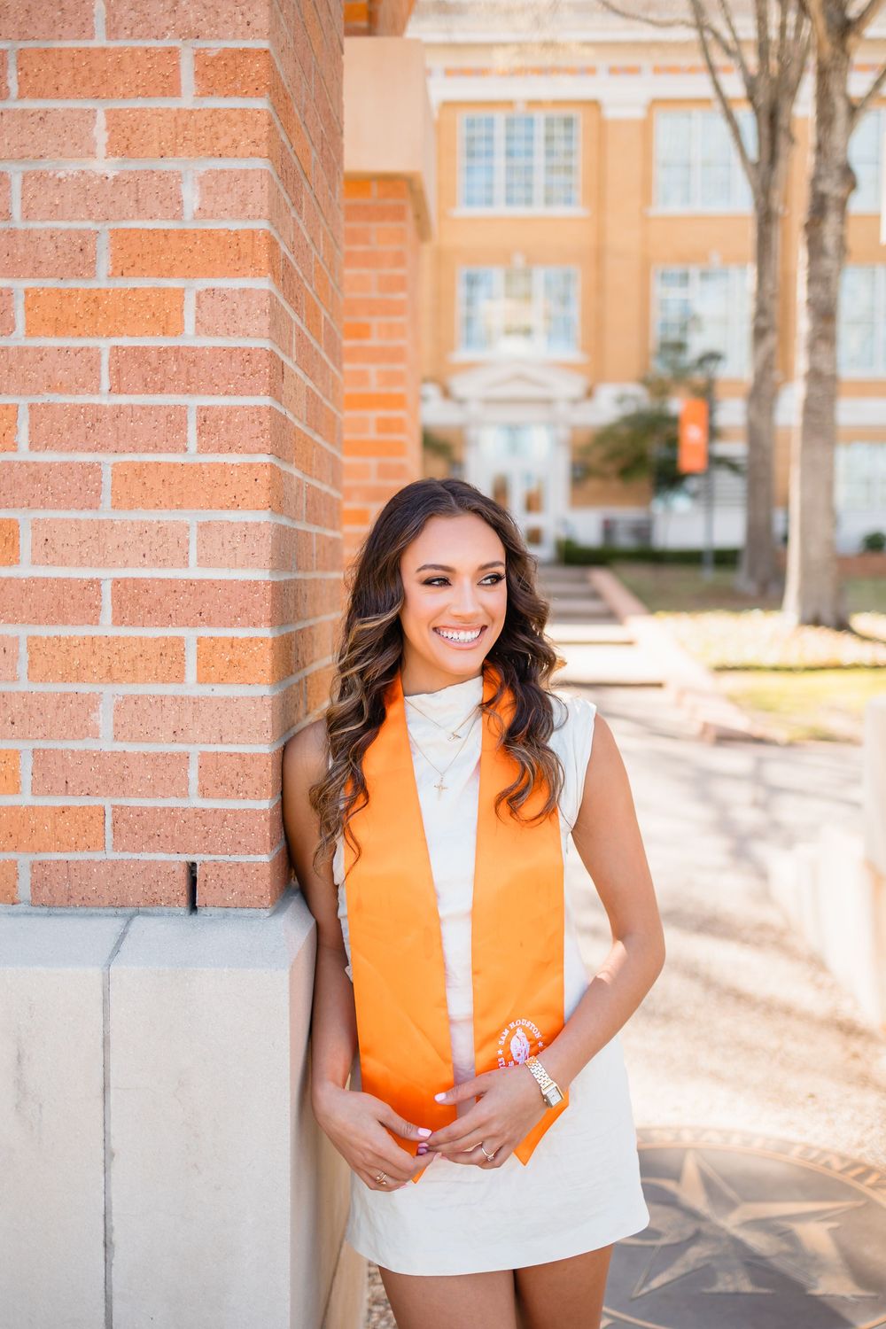 Smiling graduate in white dress with orange stole leans against brick column on sunny campus.