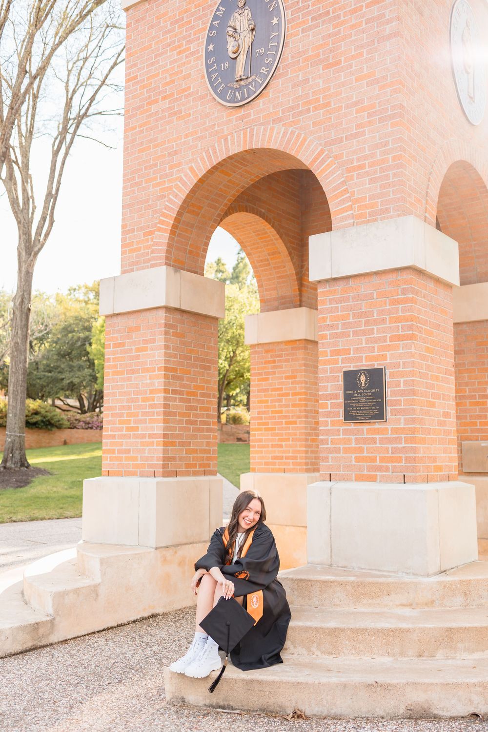 Graduate in black cap and gown sits at base of brick arch monument on university campus.