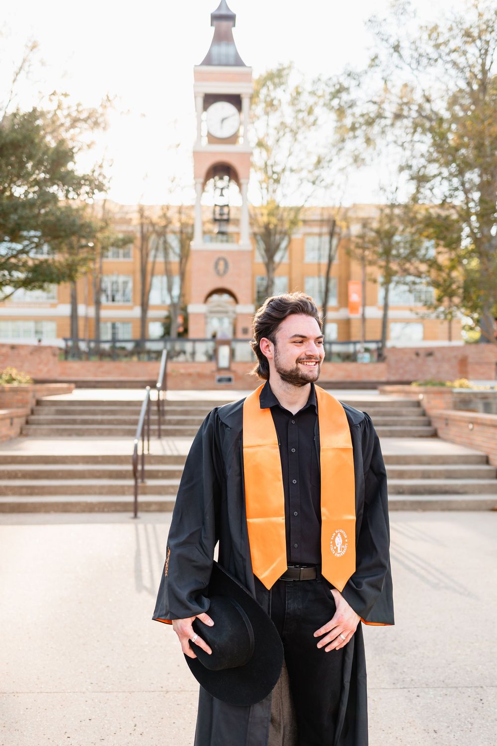 Male graduate in black cap and gown with orange stole stands before university clock tower.