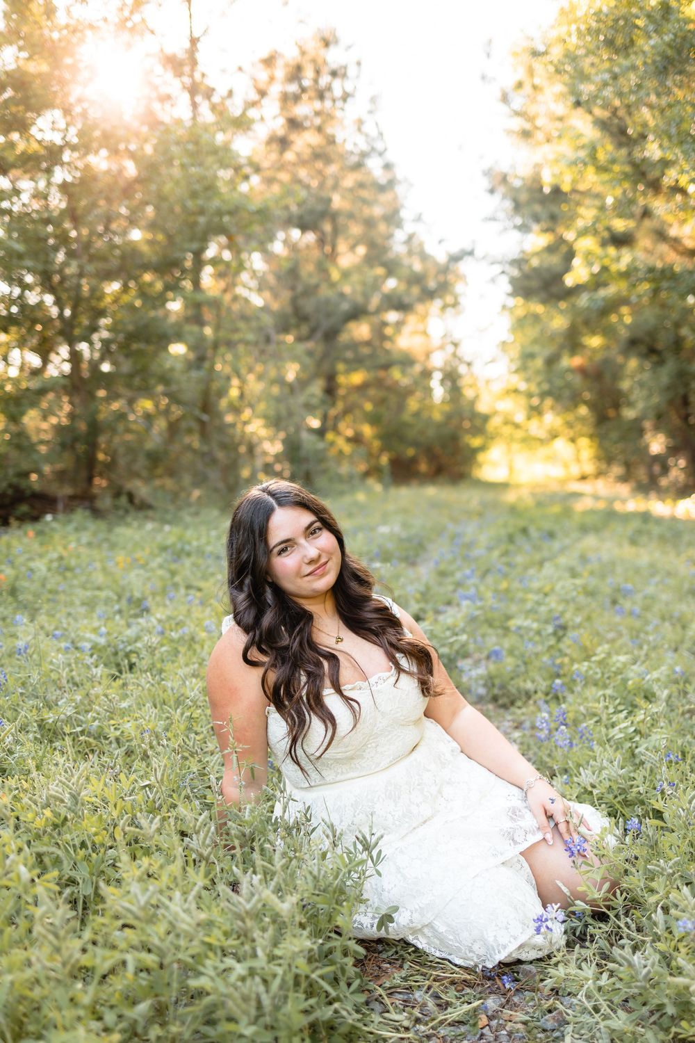 Young woman in white dress sits in sunlit field of wildflowers with golden hour lighting.