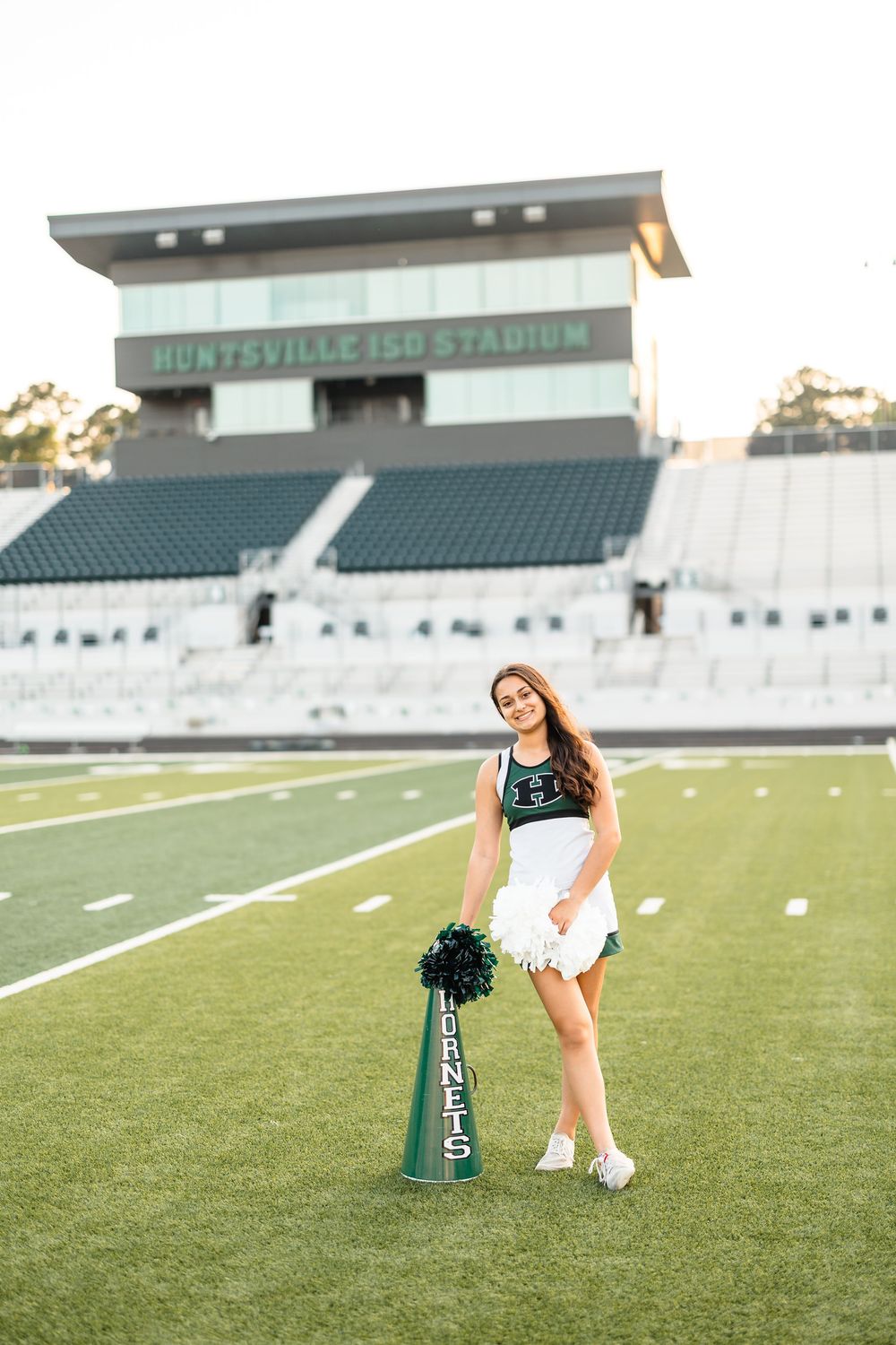 A cheerleader in uniform poses on a football field at Huntsville ISD Stadium holding a green megaphone.